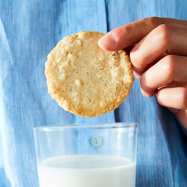 Hand holding a round, golden-brown oat cookie above a glass of white milk. Blue denim background.