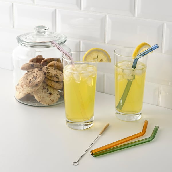 Two glasses with lemon drinks, colourful glass straws, and a cookie jar on a tiled surface.