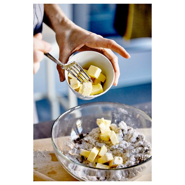 A person adds butter from a bowl to a mixing bowl with flour. Clear glass bowls on a wooden surface.