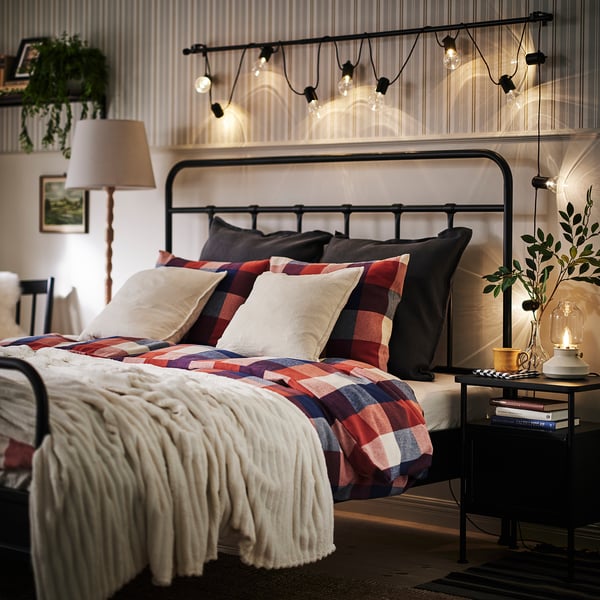 Bedroom with BLÅSENAP red and navy chequered bedding in soft cotton flannel, highlighted by warm string lights, creating a cosy atmosphere.