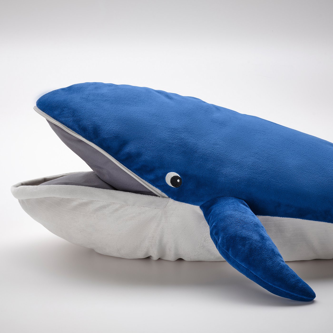 Blue whale cuddly toy, soft, detailed stitching, one visible eye, grey pillow underneath, realistic tail fin.