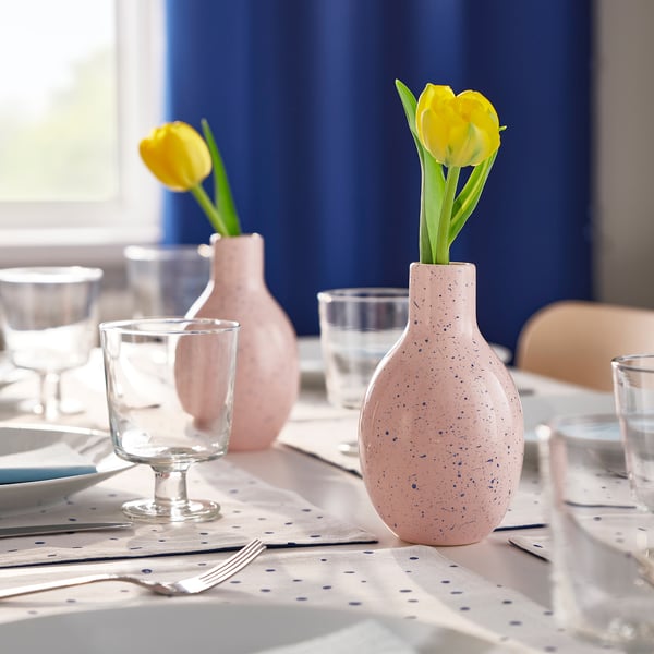 Two pink vases with blue dots on a tablecloth, holding yellow tulips alongside glassware and utensils.