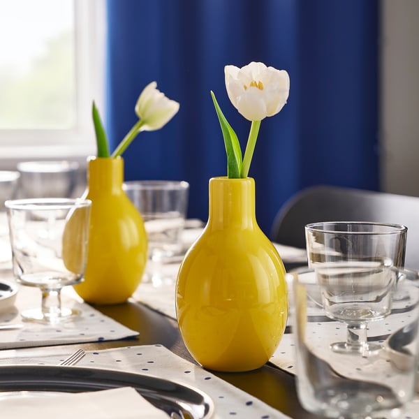 Three yellow vases hold single white flowers on a table set with glassware and blue polka dot napkins.