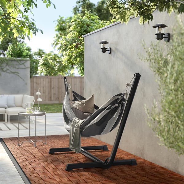 Modern outdoor hammock chair with black stand, grey fabric, cushions, on wooden deck, next to a side table and wall lights.