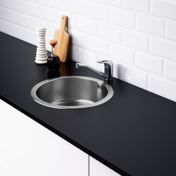 Stainless steel kitchen sink, chrome tap, black counter, white tiles, cutting board, condiments.