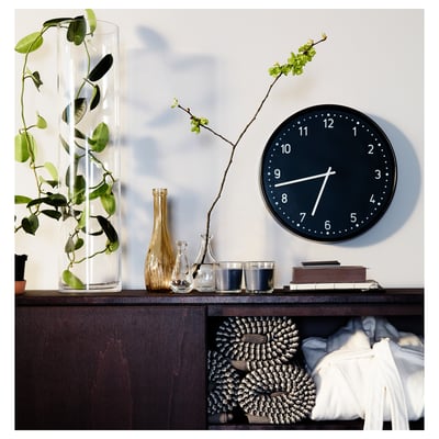 A sleek black wall clock with white numbers and hands, silent quartz movement, mounted above a dark wooden shelf with plants, vases, and candles.