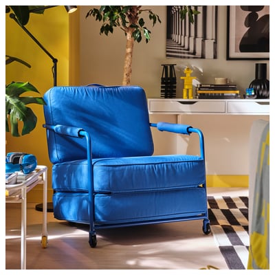 Bright blue BRÄNNBOLL chair with arms, on wheels, sitting near white side table with gaming gear. Modern design, padded, in bright room with plants.