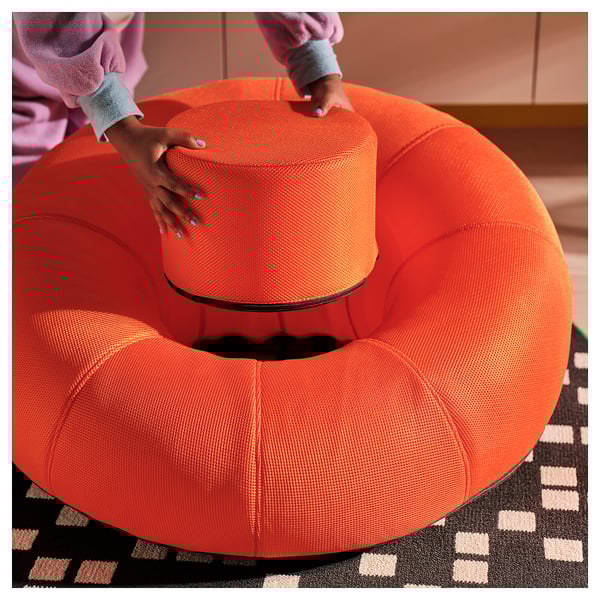 A person inflates a bright orange, mesh-like BRÄNNBOLL lounge chair for comfortable seating, shown on a geometric rug, with an included pump.