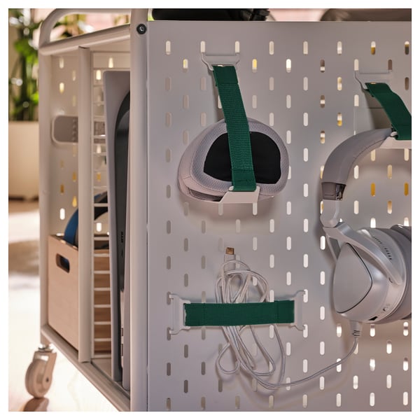 White gaming storage unit with pegboard, holding headphones, straps, and cables. Wheels for mobility, designed for organising gaming gear.