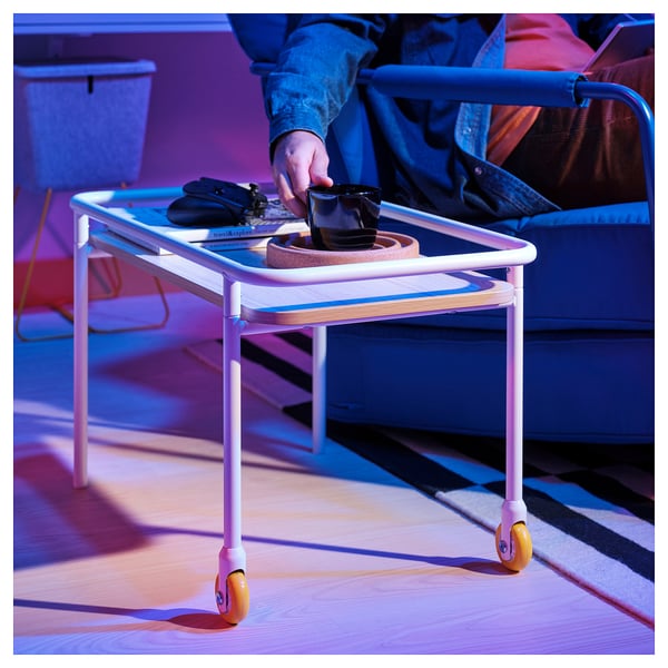 White side table with orange wheels, holding tray, phone, and coffee cup. Persons hand reaches for the cup. Ideal for snacks and gaming setup.
