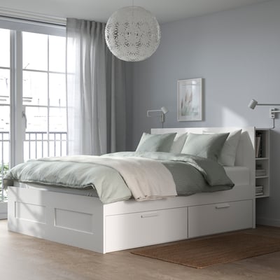 BRIMNES white bed frame with storage, neatly made with light blue linens, large window with sheer curtains and round light fixture above.