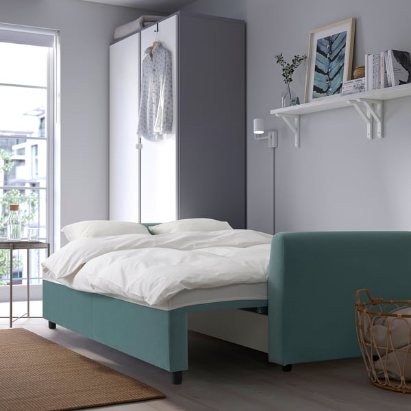 Modern bedroom with BRISSUND teal sleeper sofa, white bedding, and wall shelf, near window.