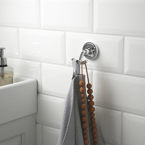 A chrome double hook on a tiled bathroom wall holds a grey towel and a wooden bead string, near a white sink with soap dispenser.