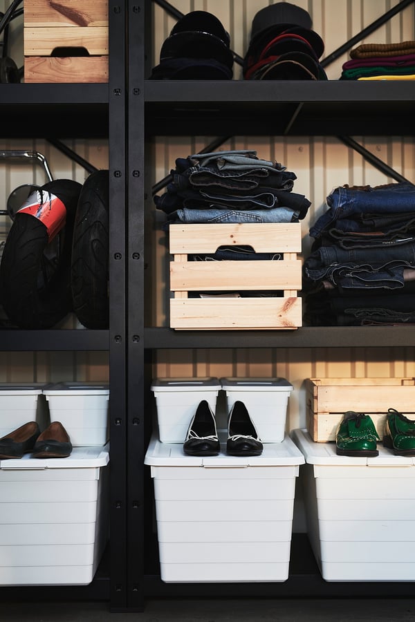 A black metal storage system, BROR, with organised shelves. It holds white bins, folded clothes, and wooden crates.