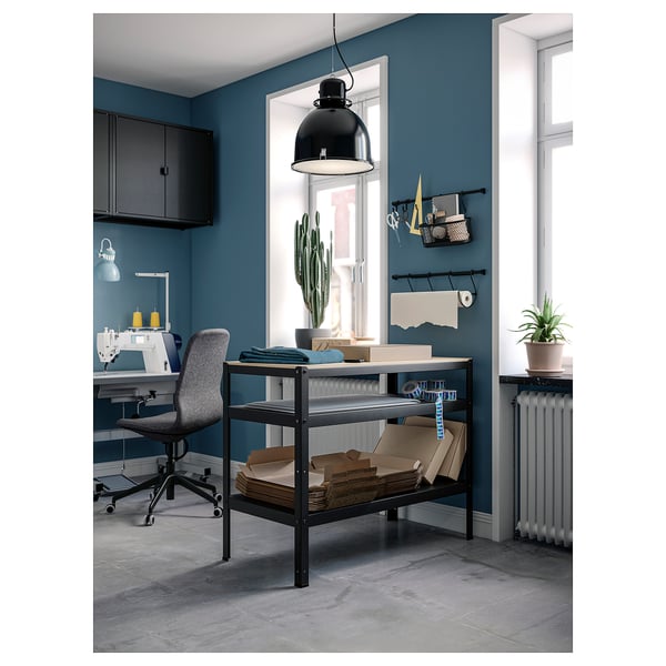 Home office with blue walls, a black metal workbench, wooden tabletop, grey office chair, sewing machine, potted plants, and a black pendant lamp.
