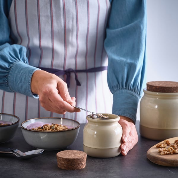 A person pours granola from a rustic stoneware jar, displaying its functional use for food storage.