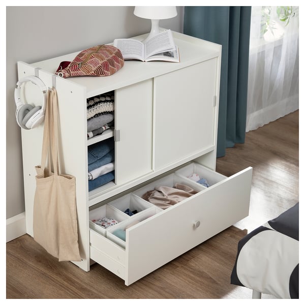 White BRUKSVARA storage unit with open drawer, folded clothes, headphones, and books on top.