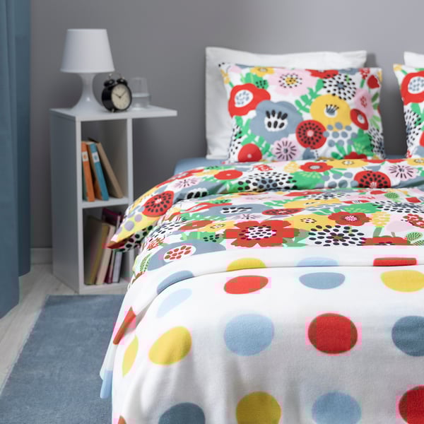 Colourful floral bedding, white pillows, bedside table with lamp, books, and clock.