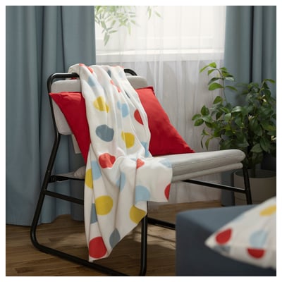 This image shows a white throw blanket with multicoloured dots draped over a grey chair with red pillows, near a window with blue curtains.