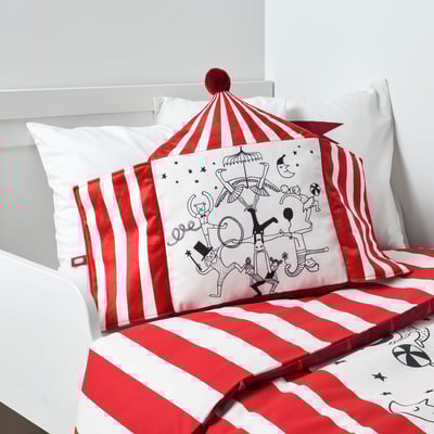 Circus-themed cushion on striped bed, supporting tired heads, made from recycled polyester.