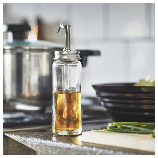 Clear glass oil dispenser with metallic nozzle on kitchen counter near chopping board and green onions. Simple, modern design.