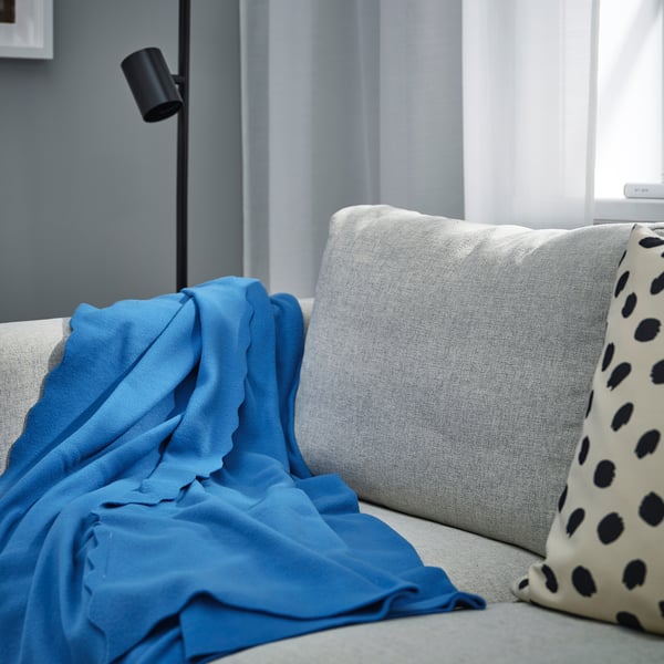A cosy blue DAGGKÅPOR fleece throw, predominantly covering a light grey couch, with black-polka dot pillows and a lamp nearby.