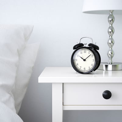 Black alarm clock on white bedside table, displaying time 12:30, beside a white pillow and lamp.