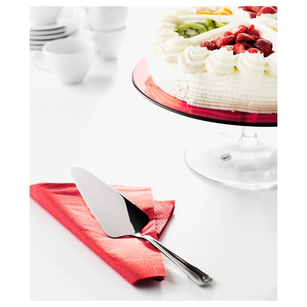 A cake on a glass stand, with whipped cream and fruit toppings. Below, a cake slicer rests on a red napkin, ready for serving.