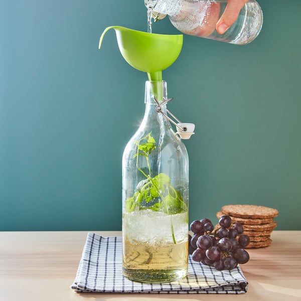 Hand pours liquid into a bottle with herbs using a funnel. Bottle on chequered cloth with grapes and cookies on a table.