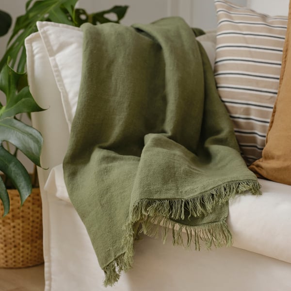 Olive green throw blanket with fringes on beige couch, striped pillow.