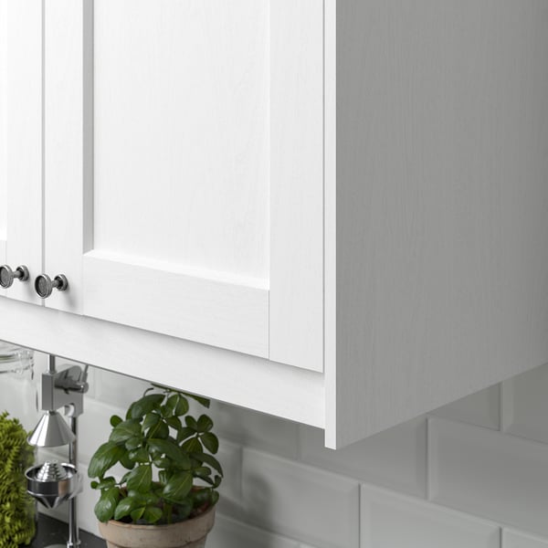 White kitchen cabinet with subtle wood grain texture. Underneath, a green potted plant sits on the counter, adding a touch of nature.