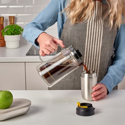 A person pours coffee from a clear pitcher into a stainless steel ENVÄLDIG travel mug with a black lid on a kitchen counter.