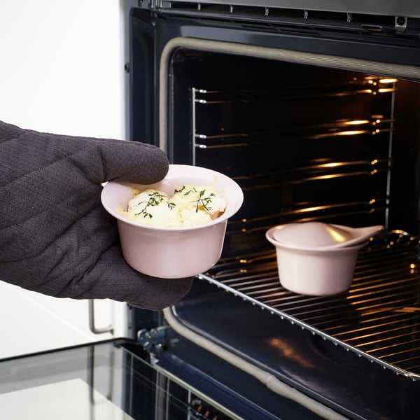Hand holding small pink oven dish with food near open oven.