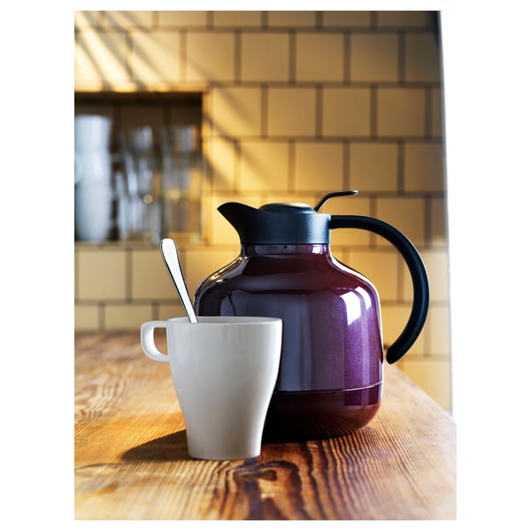A red teapot with black handle and lid, sitting on a wooden table beside a white mug with a spoon, against a tiled wall.