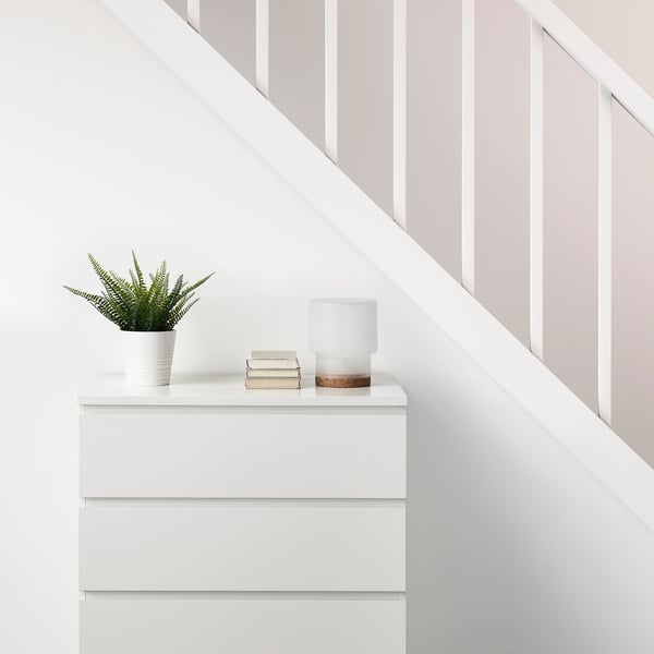 White chest of drawers with a FEJKA artificial plant, stack of books, and white lamp on top, next to a staircase.