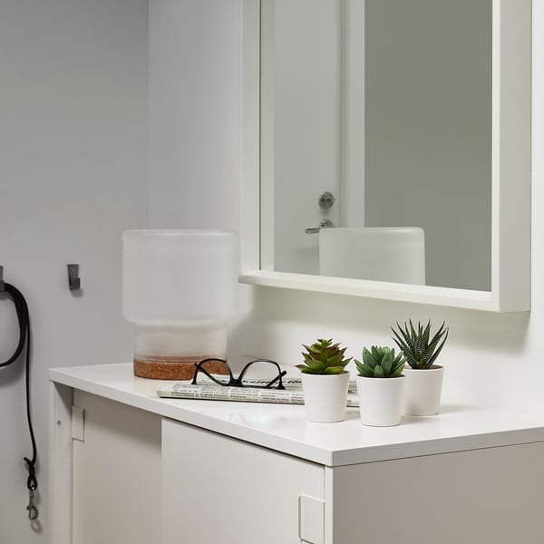 Modern bathroom counter with white cabinet, mirror, FEJKA artificial plants, and lamp. Laundry hook and book also.