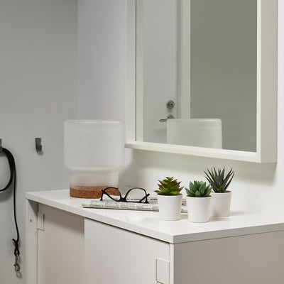 Modern bathroom counter with white cabinet, mirror, FEJKA artificial plants, and lamp. Laundry hook and book also.