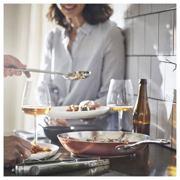 Kitchen: person serving food from copper pan on dark counter, wine, plates.