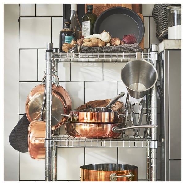 A multi-tiered kitchen shelf organiser holds copper pots, a stainless-steel funnel, fresh vegetables, and wine bottles.