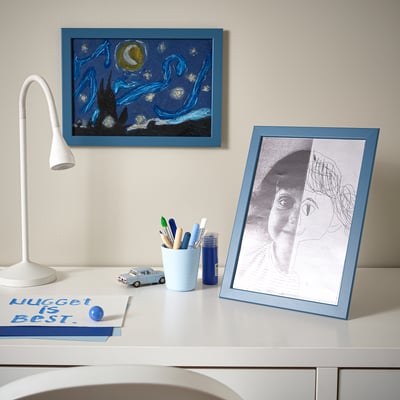 A desk with FISKBO frames; one shows a childs drawing, the other a night sky. Stationery, a lamp, and a book are neatly arranged.