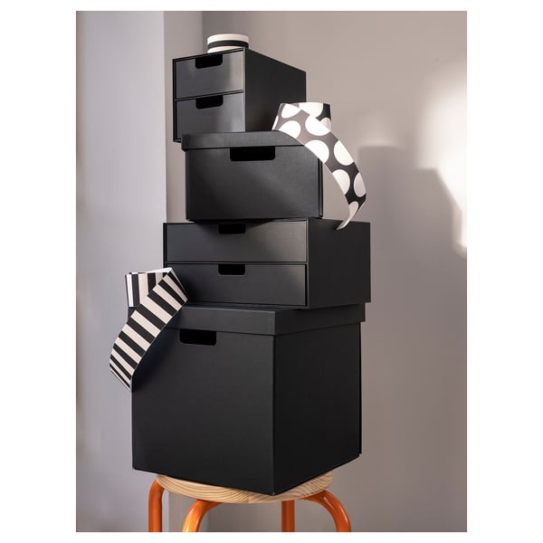 Stack of black FJÄDERHARV boxes on an orange stool. Boxes are square with cut-out handles. One box has a striped design, another has polka dots.