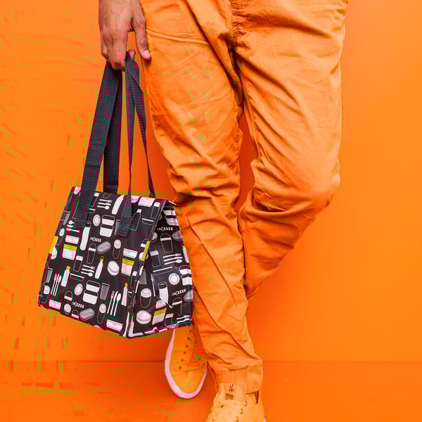 Person holds black patterned lunch bag against orange background. Bag playful designs of mugs and bottles.