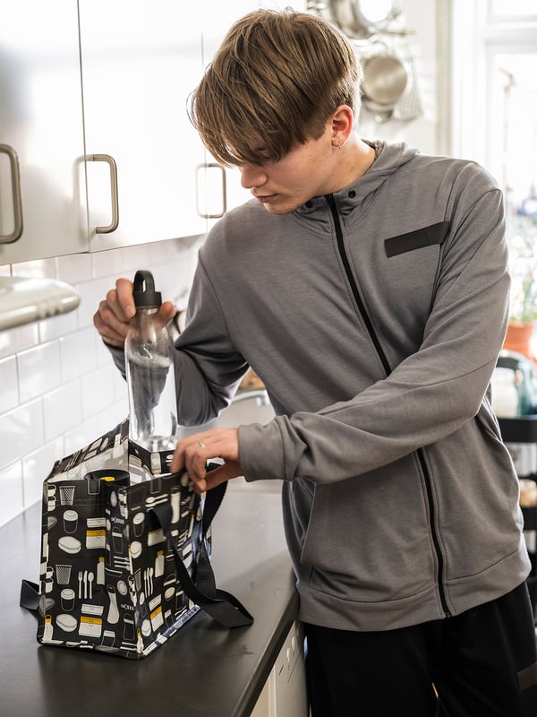 Person in grey jacket packs clear bottle into black FLADDRIG lunch bag with kitchen tools pattern in white kitchen.