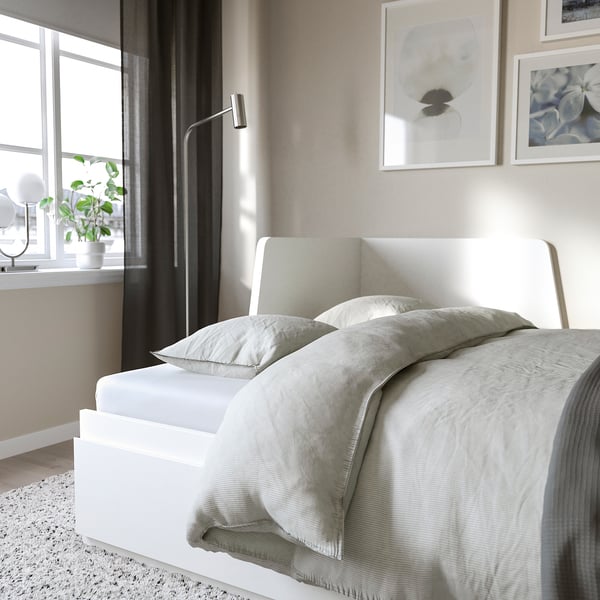 Modern bed with white headboard, light grey bedding, under-window houseplant, and wall art in minimalist bedroom.