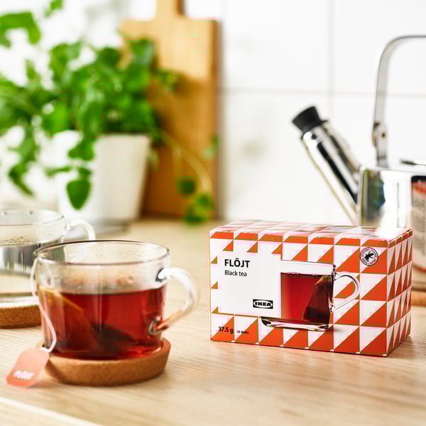 Clear mug of steaming black tea beside an opened ikea FLÖJT tea box with 25 tea bags on a wooden table, suggesting its ready to drink.