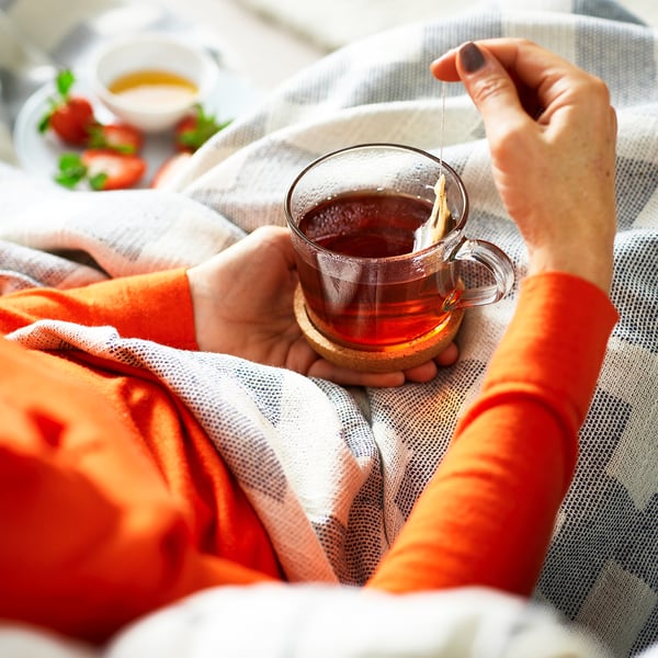 Person holding a clear glass mug with a tea bag, surrounded by cosy blankets and a bowl of strawberries.