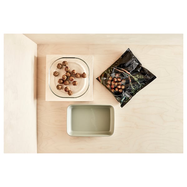 A clear glass oven dish, square-shaped, holding food. Its placed on a wooden surface with a green rectangular tray and a packaged food item nearby.