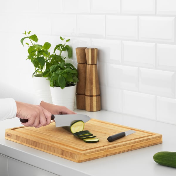 Person slicing cucumber on wooden cutting board with two knives beside potted herbs and wooden mills.