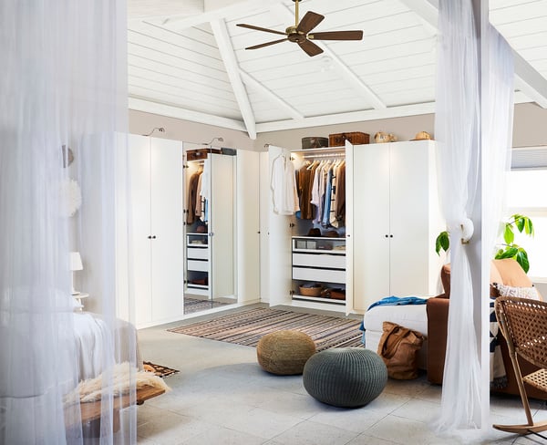 Bright, minimalist bedroom white wardrobes, a ceiling fan, plush fabric pouffes, and sheer white curtains creating a serene atmosphere.