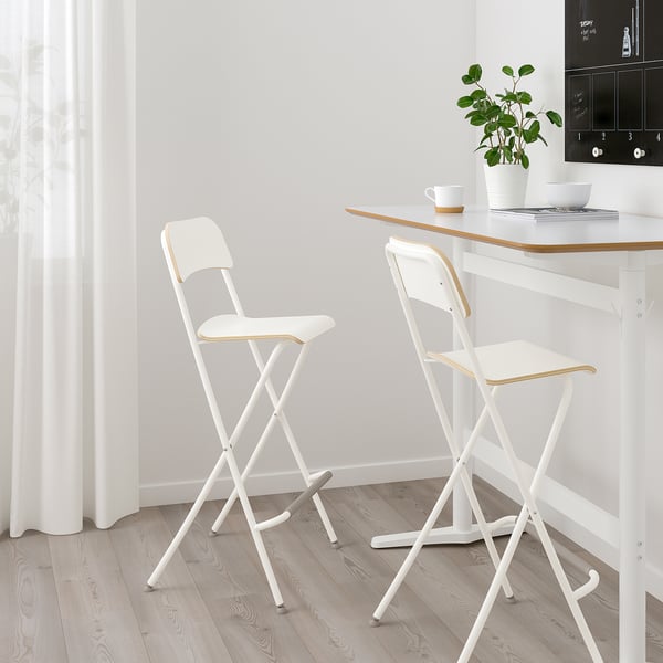 Two white FRANKLIN foldable bar stools by wooden table, simple, modern design.